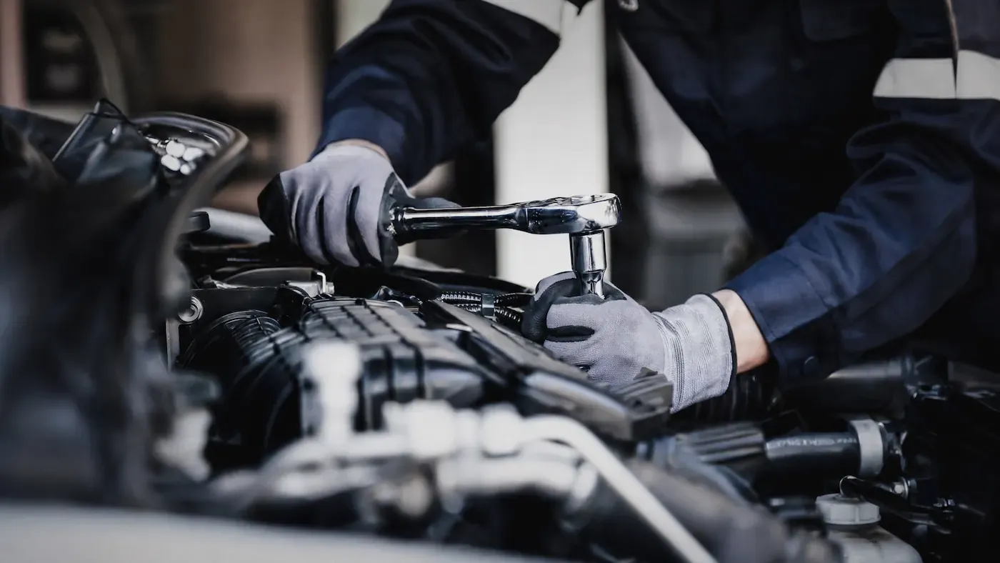 Mechanic in a navy coverall and grey gloves using a ratchet on an engine under an open car hood.