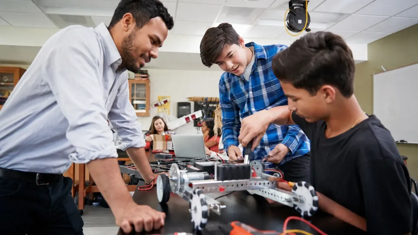Instructor and two students assembling a wheeled robot at a classroom table, with classmates at a laptop in the background.