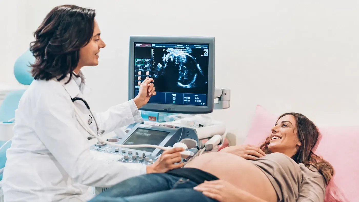 Clinician in a white coat performing an ultrasound on a smiling pregnant patient beside a monitor showing the scan.