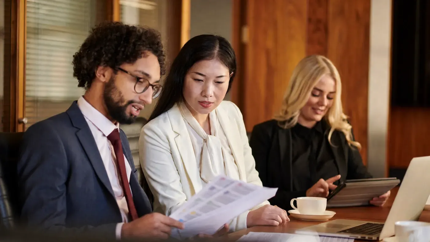 Three colleagues in business attire reviewing a printed document and a tablet at a conference table.