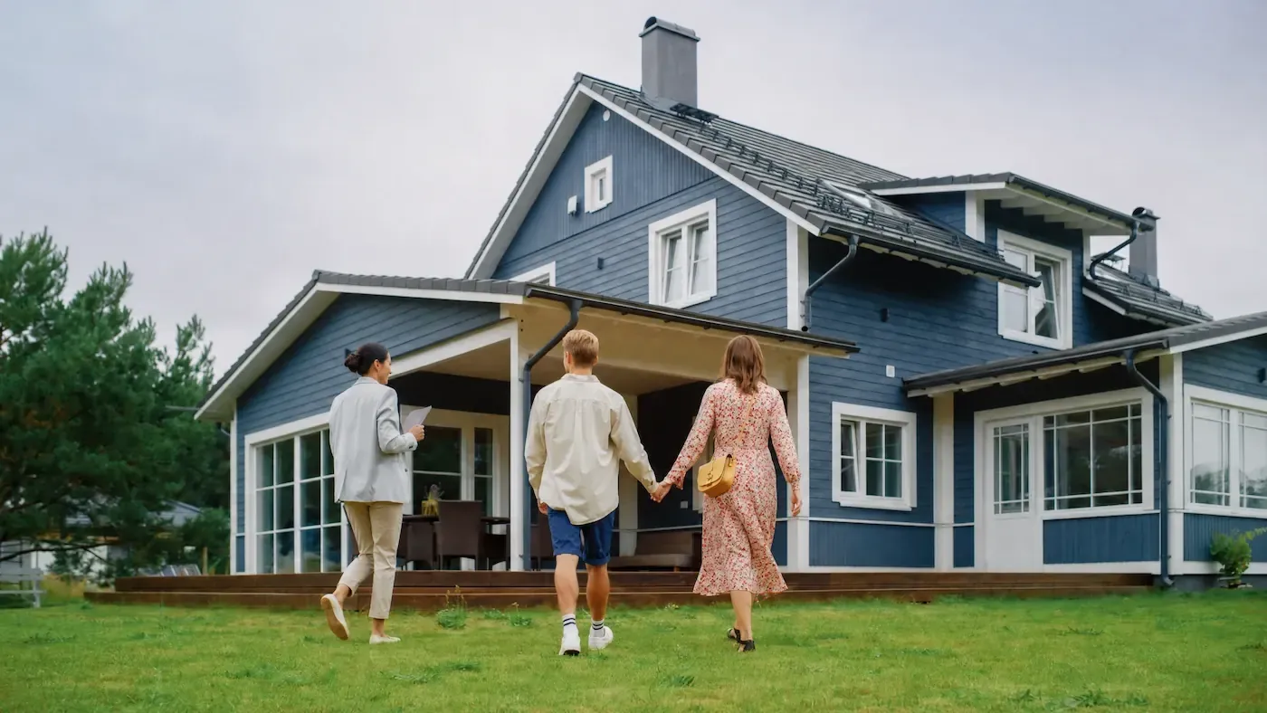 Couple and an agent walking toward a blue two-story house with white trim across a green lawn.