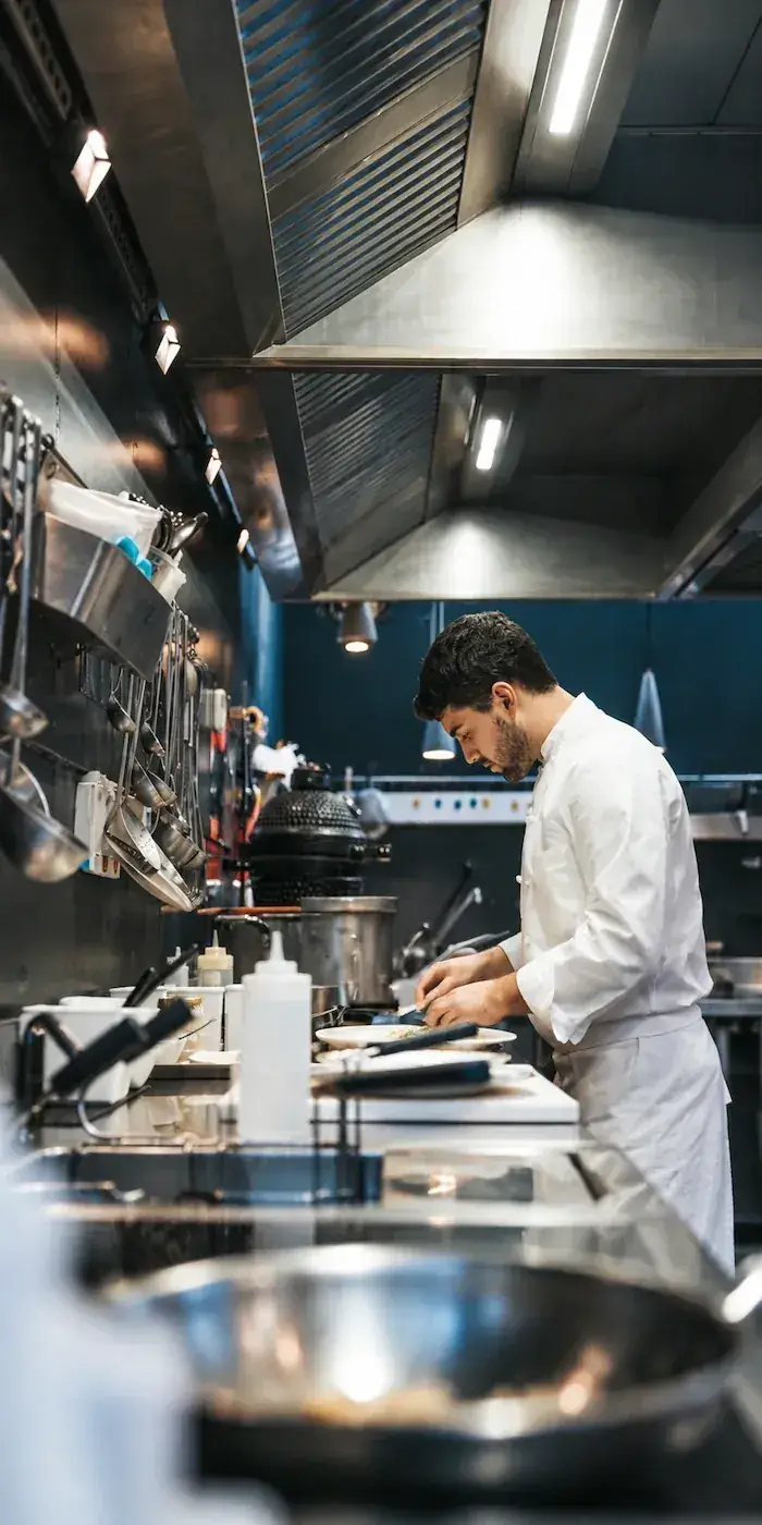 Chef in a white coat plating a dish at a stainless-steel restaurant pass.