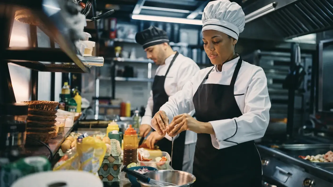 Chef in a white coat, black apron, and tall toque cracking an egg at a stainless prep line, with another chef working behind her.