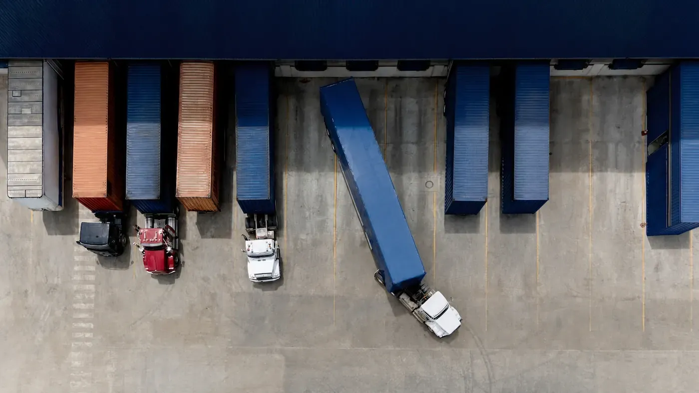 Overhead view of tractor-trailers parked at a warehouse loading dock, with one blue trailer angled into the yard.